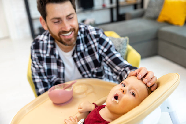 Bearded adult father feeding his infant baby in home interior teaching toddler kid eating from spoon. - Фото, изображение