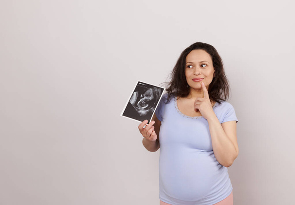 Mulher grávida pensativa latino-americana emocional, segurando ultra-som de seu bebê, sonhadamente olhando para o lado um espaço de cópia no fundo branco. Conceito de diagnóstico de gravidez. Obstetrícia e ginecologia - Foto, Imagem