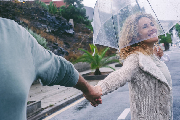 Ευτυχισμένο ζευγάρι απολαμβάνουν χρόνο και υπαίθρια δραστηριότητα αναψυχής μαζί κάτω από τη βροχή χρησιμοποιώντας ομπρέλα. Ο Ποβ άντρας κρατάει τη γυναίκα χέρι-χέρι. Ευτυχία και γαλήνια ζωή. Άνθρωποι περπατούν στο δρόμο - Φωτογραφία, εικόνα