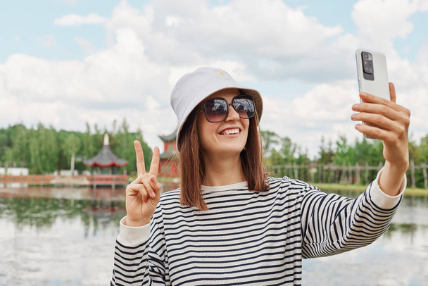 Smiling happy woman wearing striped shirt panama sunglasses standing near lake making selfie showing v sign having video call traveling enjoying sightseeing. - Foto, afbeelding