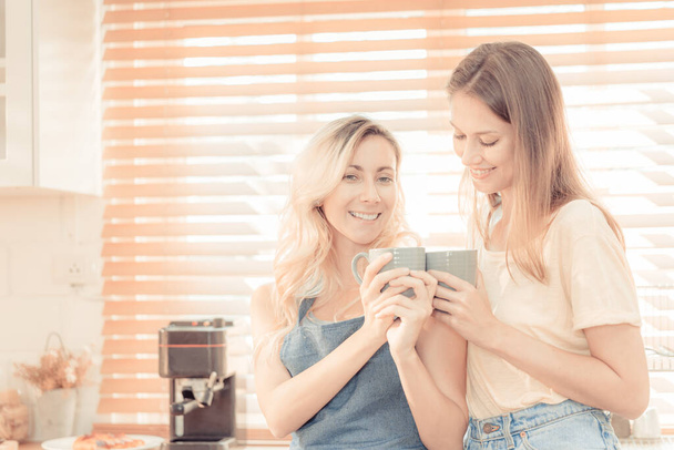 happy lesbian couple holding cups of coffee in kitchen. Couple of lesbian Girls Enjoy coffee at home taking about something. Two young adult beautiful women drink tea in modern kitchen. warm tone. - Fotoğraf, Görsel