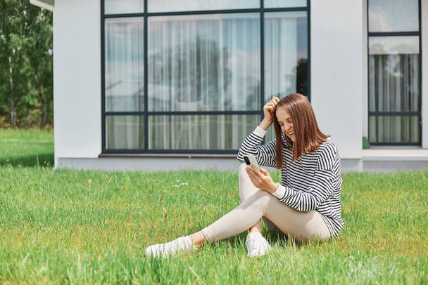 Attractive happy woman wearing striped shirt and leggins sitting on grass against modern house with big windows using her mobile phone for browsing internet or having video call. - Foto, Bild