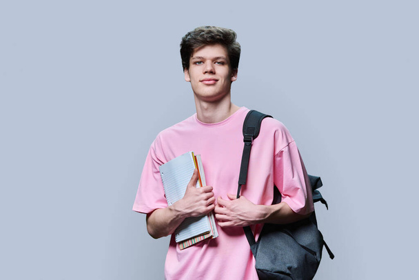 Young guy college student with backpack holding notebooks textbooks, in pink t-shirt looking at camera on grey studio background. Education, studying, learning concept - Foto, Imagem