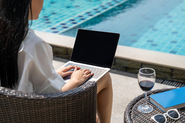 Młoda Azjatka z długimi czarnymi włosami używa laptopa ze szklanką czerwonego wina. Siedzi przy basenie.  - Zdjęcie, obraz