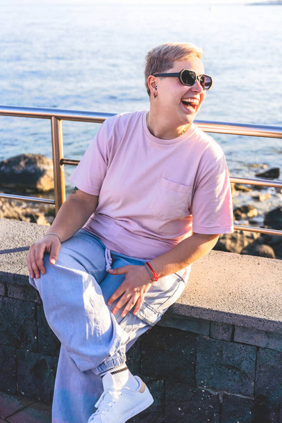 Androgyne nichtbinäre Person mit lebhaften kurzen Haaren, Piercings und sechseckiger Sonnenbrille, die am Meer lacht. Sommerstimmung und schöne Küste - Foto, Bild