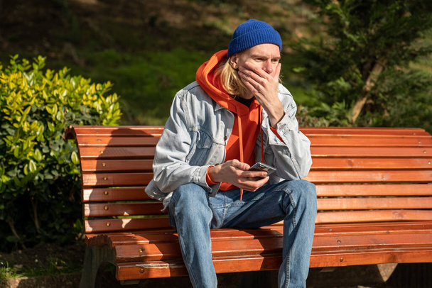 Aufgebrachter europäischer Hipster-Mann, der mit geschlossenem Mund auf einer Bank sitzt und sein Mobiltelefon in der Hand hält und schockiert auf die schlechte Nachricht blickt. Frustrierte Skandinavier erhalten traurige Nachricht. Männer brauchen psychologische Hilfe - Foto, Bild