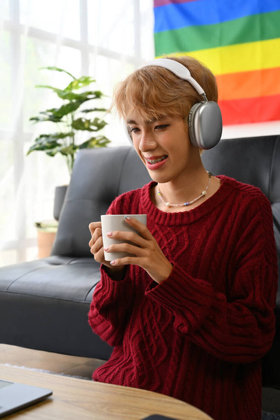 Bastante joven gay hombre usando auriculares beber café viendo vídeos, navegar por Internet en el ordenador portátil en casa. - Foto, imagen