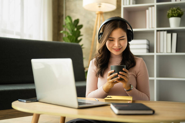 Beautiful young asian woman in headphone holding mug of coffee and using laptop at home. People, technology and lifestyle. - Foto, Imagem