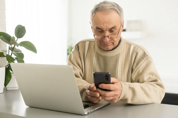 Schöner lächelnder älterer Mann mit Brille und Handy am gemütlichen Arbeitsplatz mit Laptop zu Hause, Rentner im Chat mit Freunden in den sozialen Medien, Tippen auf dem Smartphone - Foto, Bild