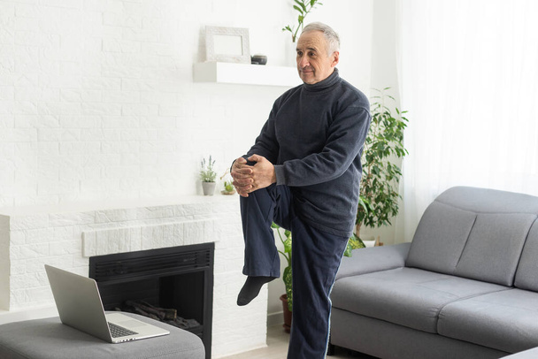 Sport in mature age. Happy middle-aged man doing stretching exercises in front of laptop at home, watching online tutorials, in living room interior. - Photo, Image