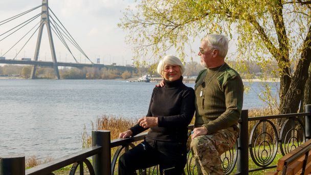 高齢者の白髪のカップル、軍事化されたカーキの服の男、黒いセーターの女性、秋には川の堤防の街での肖像画 - 写真・画像