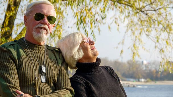 高齢者の白髪のカップル、軍事化されたカーキの服の男、黒いセーターの女性、秋には川の堤防の街での肖像画 - 写真・画像