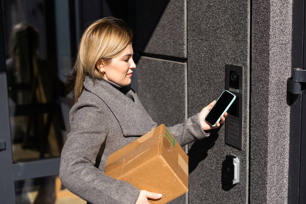 Pensive ξανθιά γυναίκα ανοιχτή πόρτα χρησιμοποιώντας smartphone με εφαρμογή mock up οθόνη, θηλυκό ξεκλειδώνει το σύστημα ασφαλείας μέσω κινητού τηλεφώνου τον κωδικό ελέγχου για την είσοδο. - Φωτογραφία, εικόνα