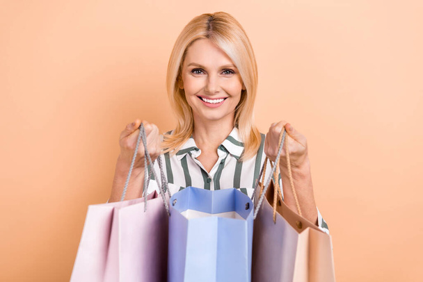 Portrait of satisfied retired woman bob hairdo striped formalwear showing new outfit in shopping bags isolated on beige color background. - Fotografie, Obrázek