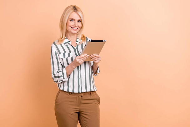 Portrait of positive optimistic retired woman wear striped formalwear hold tablet reads statistics isolated on beige color background. - Foto, Imagen