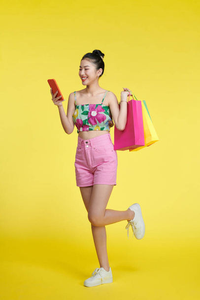 Ευτυχισμένη νεαρή Ασιάτισσα γιορτάζει με κινητό τηλέφωνο και κρατώντας τσάντες για ψώνια - Φωτογραφία, εικόνα