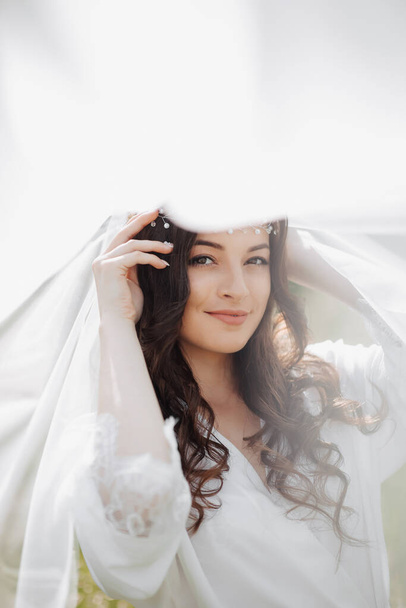 A beautiful bride with long curly hair and a delicate tiara on her head, wrapped in a white veil, gently holds her hair. Portrait of a girl looking into the lens. Natural makeup - Фото, изображение