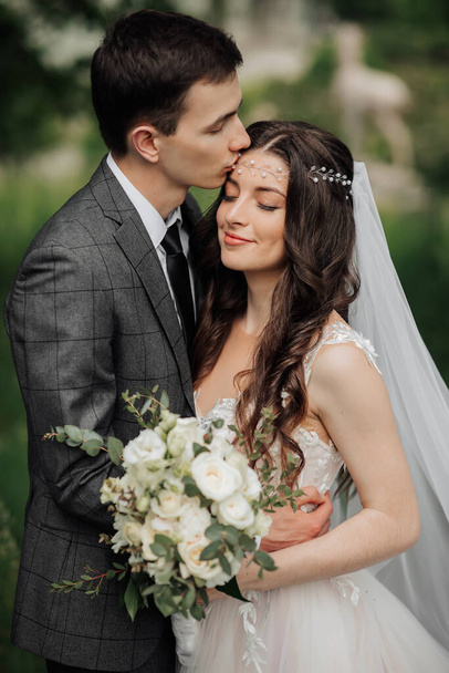 Happy young couple. Wedding portrait. The groom kisses the bride on the temple. Young people are tenderly hugging while looking at the camera. Wedding bouquet. Spring wedding - Fotografie, Obrázek