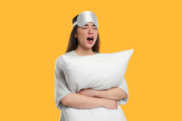 Tired young woman with sleep mask and pillow yawning on yellow background. Insomnia problem - Foto, afbeelding