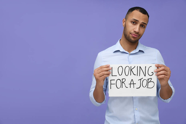 Young unemployed man holding sign with phrase Looking For A Job on violet background. Space for text - Foto, Imagem