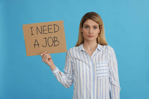 Unemployed woman holding sign with phrase I Need A job on light blue background - Fotoğraf, Görsel