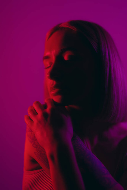 Hayalperest kadın model, neon aydınlatmada menekşe arkaplanına karşı iyilik için dua ederken gözlerini kapatıyor. - Fotoğraf, Görsel
