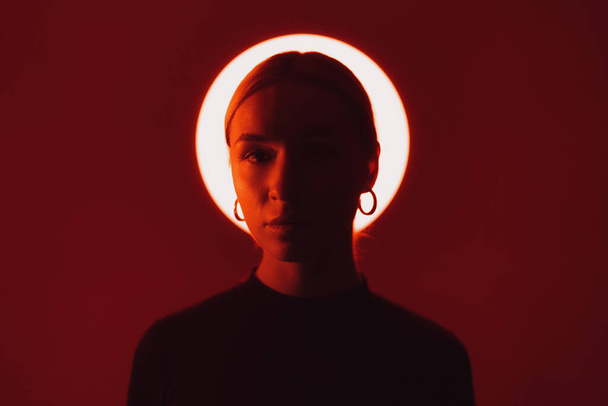 Retrato de mujer tranquila mirando a la cámara contra la luz brillante en estudio oscuro con iluminación roja - Foto, imagen
