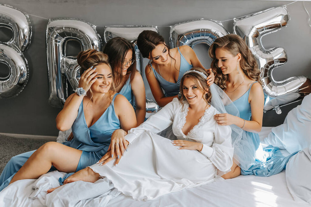 The bridesmaids look at the smiling bride. The bride and her cheerful friends are celebrating a bachelorette party on the bed in identical dresses. Bride and friends in the room - Fotó, kép