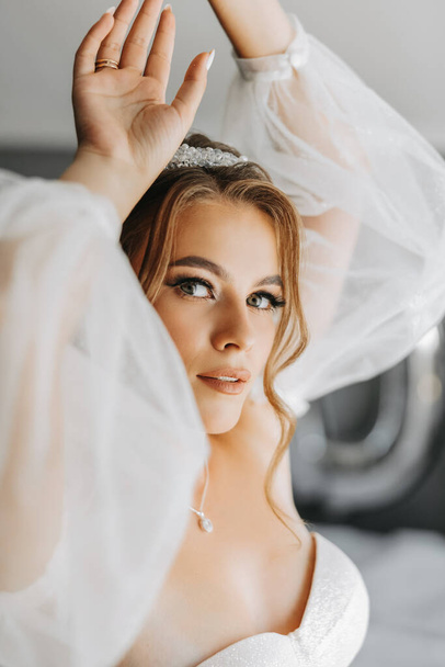 A beautiful curly brown haired bride in a white dress poses for a photographer while standing in a room in a beautiful dress with sleeves. Wedding photography, portrait close-up. - Photo, image