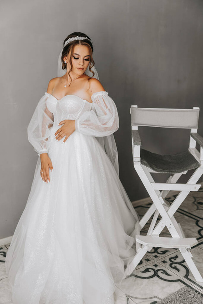 A beautiful curly brown haired bride in a white dress poses for a photographer while standing in a room in a beautiful dress with sleeves. Wedding photography, close-up portrait, chic hairstyle. - Foto, immagini