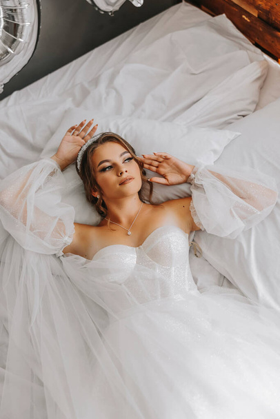 A beautiful curly brown haired bride in a white dress poses for a photographer while lying on a bed in a beautiful dress with sleeves. Wedding photography, close-up portrait, chic hairstyle. - Fotoğraf, Görsel