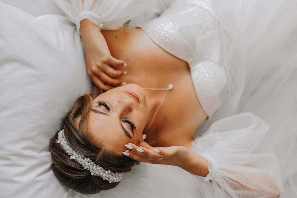 A beautiful curly brown haired bride in a white dress poses for a photographer while lying on a bed in a beautiful dress with sleeves. Wedding photography, close-up portrait, chic hairstyle. - Zdjęcie, obraz