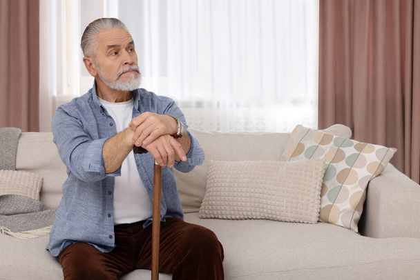 Senior man with walking cane sitting on sofa at home. Space for text - Fotó, kép