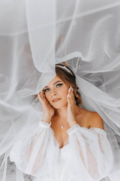 Beautiful curly brown-haired bride in a white dress poses for a photographer, standing under a veil in a beautiful dress with sleeves. Wedding photography, close-up portrait, chic hairstyle. - Photo, Image
