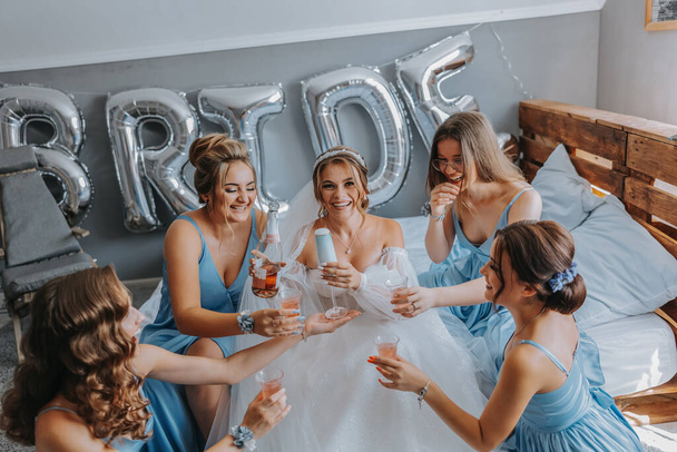 Young bridesmaids in blue silk dresses drink champagne in the bride's room. Beautiful women celebrating bachelorette party sitting on bed and with champagne. - 写真・画像
