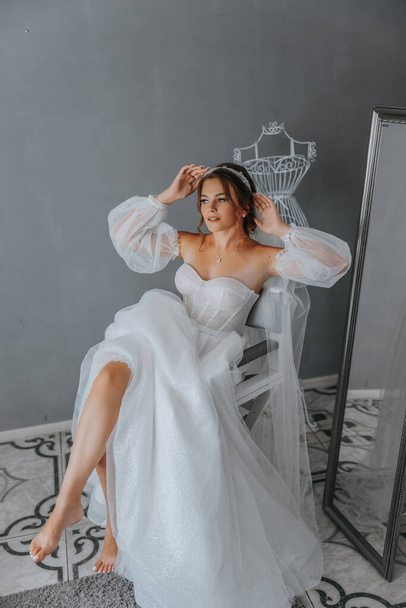 Beautiful curly brown-haired bride in a white dress poses for a photographer, standing under a veil in a beautiful dress with sleeves. Wedding photography, close-up portrait, chic hairstyle. - Фото, изображение