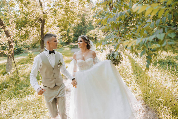 Portrait of a beautiful couple in love on your wedding day. A walk in the park in the sunlight, the groom leads the bride. Amazing kisses and hugs of the bride and groom with a bouquet - Photo, Image