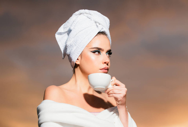 Attractive sexy woman holding a cup with coffee and looking at the sunrise. Beauty woman with towel on head smelling coffee in the night at dramatic sky outdoor. Morning spa - Fotó, kép