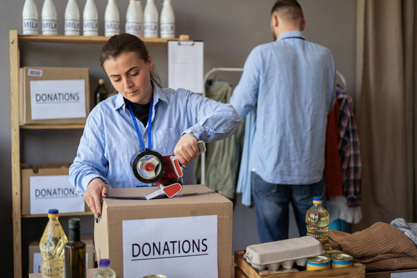 People working in charitable foundation. Female volunteer with scotch dispenser closing package near carton boxes in charity center. Volunteers sort donations during food drive. Copy space - Photo, Image