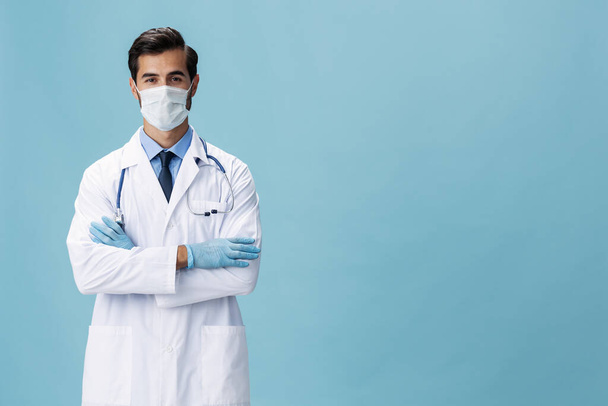 Beyaz önlüklü ve tıbbi maskeli erkek doktor el hareketlerini gösteriyor ve mavi izole arkaplandaki kameraya bakıyor, kopyalama alanı, yazı için boşluk. Yüksek kalite fotoğraf - Fotoğraf, Görsel