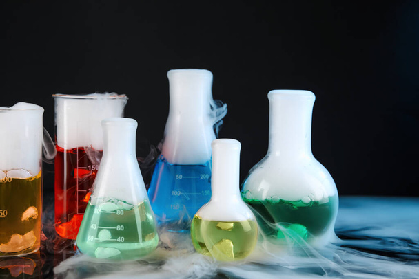 Laboratory glassware with colorful liquids and steam on black background. Chemical reaction - Photo, Image