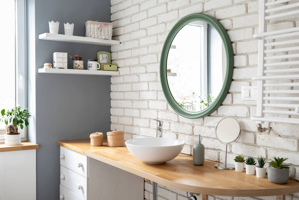 Industrial interior of bathroom with window, wooden furniture and counter with ceramic sink and round mirror on white brick wall. Modern design at home. - Foto, imagen