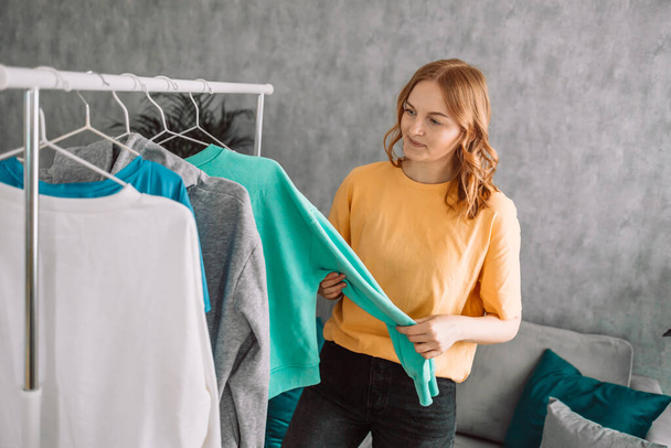 Clothing, purchasing, style concept. Happy blonde haired woman chooses clothes at home wardrobe, holds comfortable jumper on hangers, outfits on racks in background, messages via mobile phone. High - Photo, Image