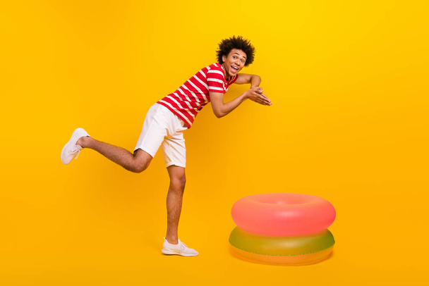 Full length photo of excited cool guy wear striped t-shirt diving inside two inflatable rings isolated yellow color background. - Fotografie, Obrázek