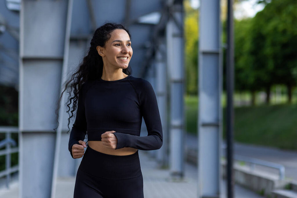 Young beautiful hispanic woman with curly hair jogging near stadium, woman in tracksuit smiling contentedly, running in park. - Foto, afbeelding
