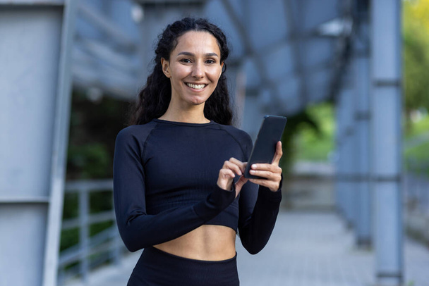 Outdoor portrait active sportswoman outdoors in the air near an urban, an athlete smiling and looking at the camera, a Latin American woman with curly hair and wearing a tracksuit is holding a phone. - Photo, image