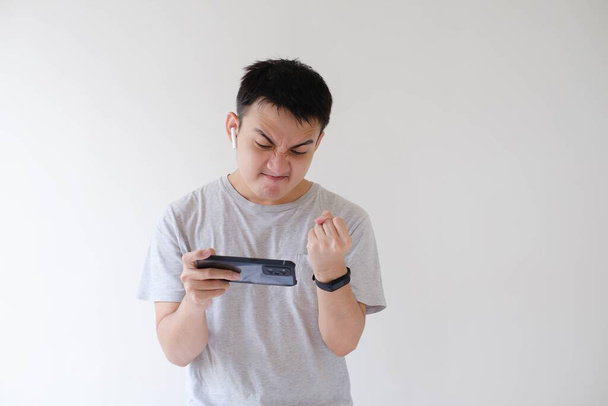 Un joven asiático que lleva una camiseta gris está enojado mientras juega un juego en su teléfono inteligente. Fondo blanco aislado. - Foto, Imagen