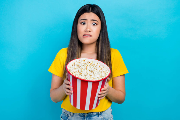 Photo of young nervous frightened girl korean hold bucket popcorn eating snacks scared thriller evil film isolated on blue color background. - Foto, imagen
