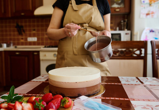 牛乳、ダークチョコレート、ホワイトチョコレートで作られた層状のおいしいケーキに焦点を当てる-キッチンテーブルのトリプルチョコレートムースデザートと釉薬でボウルを保持主婦。料理だ。菓子類 - 写真・画像
