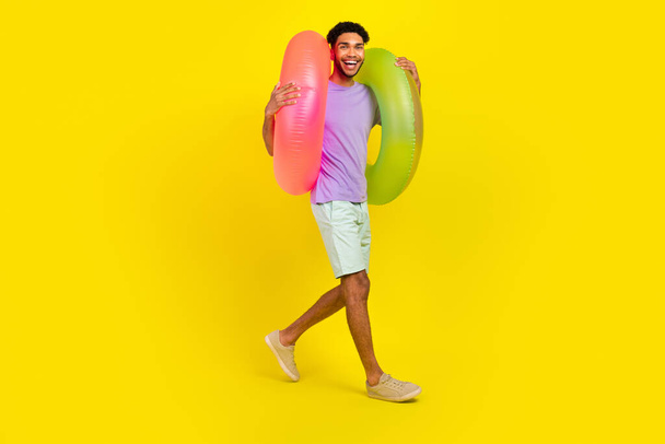 Full size body cadre of young guy enjoy pool parties walking with inflatable rings lifebuoy beach tour isolated on yellow color background. - Foto, Bild